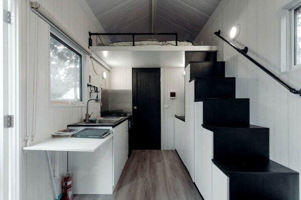 A compact kitchen with a built-in stove, a sink, and a small countertop. There is a black staircase leading to an upper level.