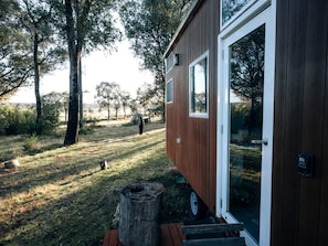A small wooden cabin with a glass door, situated in a grassy area with trees in the background.