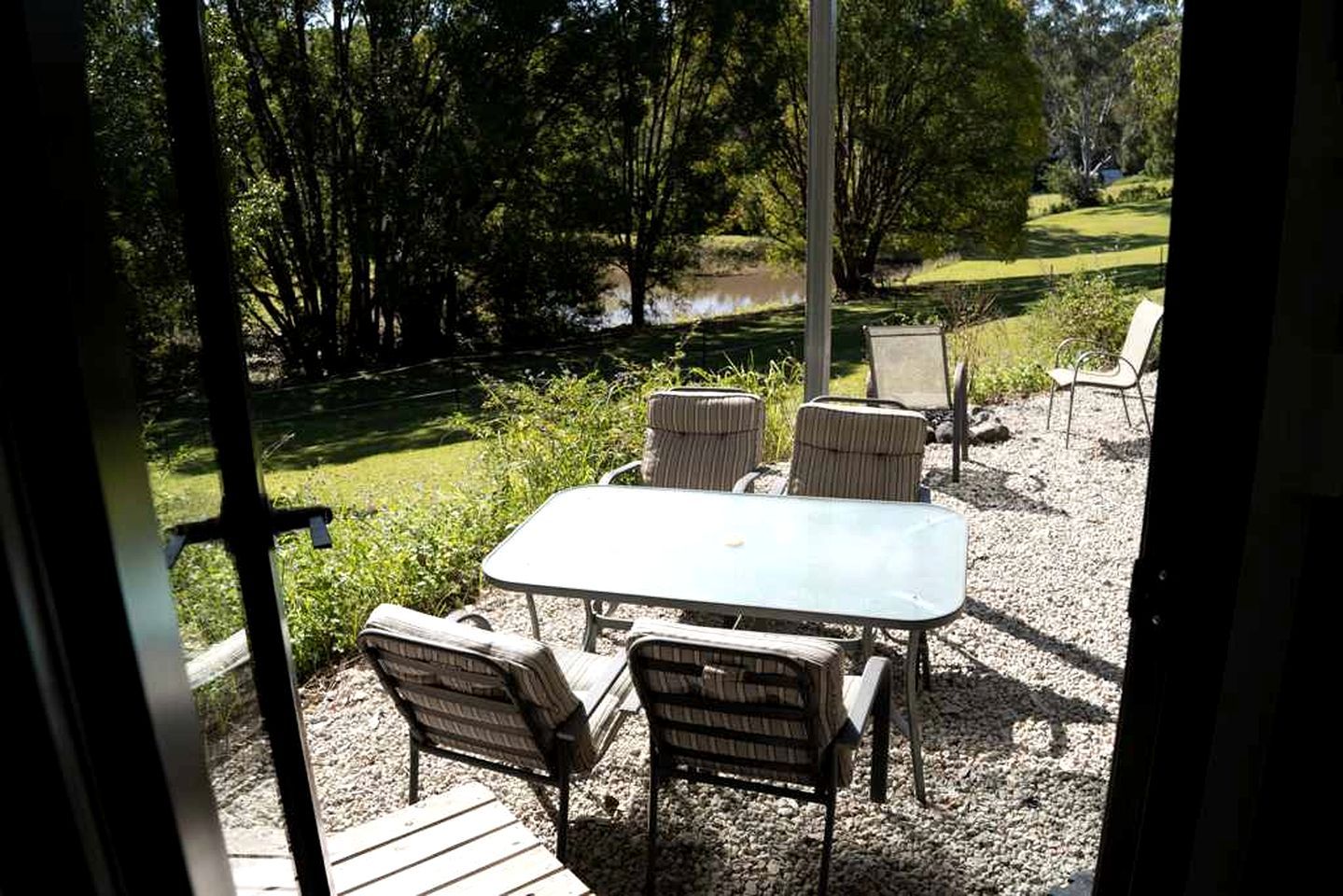 An outdoor patio with a table and chairs, surrounded by gravel and greenery, with a view of a lake and trees.