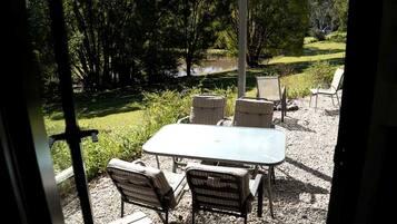 An outdoor patio with a table and chairs, surrounded by gravel and greenery, with a view of a lake and trees.