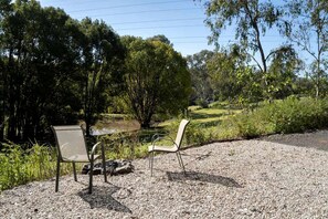 Deux chaises et une table sont placées sur une surface de gravier, offrant une vue sur une zone herbeuse et des arbres.