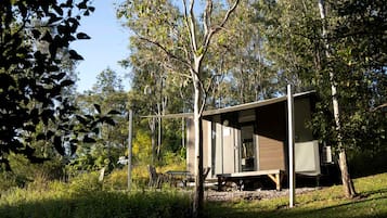 A modern, single-story cabin with a large glass door, surrounded by dense trees and natural vegetation.