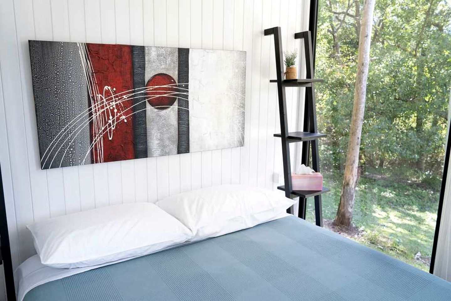 A bedroom with a bed, a wall-mounted abstract painting, a black shelving unit with a plant, and a window with a view of greenery.