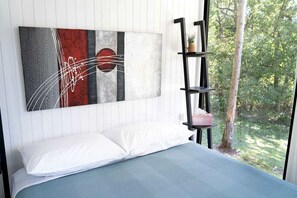 Quarto com cama, pintura abstrata na parede, estante preta com uma planta e janela com vista para vegetação.