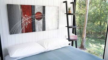 Un dormitorio con una cama, un cuadro abstracto en la pared, una estantería negra con una planta y una ventana con vistas a zonas verdes.