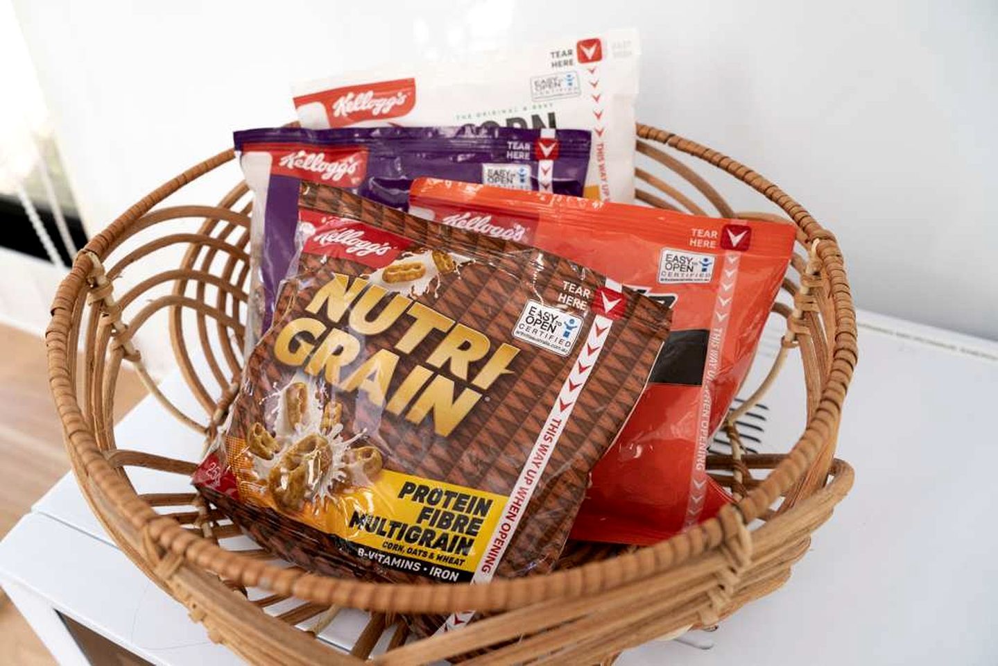 Un panier en osier contenant divers produits alimentaires emballés, notamment un paquet de céréales multigrains de la marque Nutri Grain.