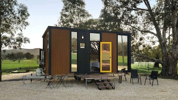 Una cabaña moderna con un amplio ventanal, mobiliario exterior y vistas del paisaje circundante.