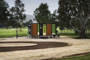 A modern RV-style accommodation with a dining area, situated in a rural setting with trees and a grassy field.