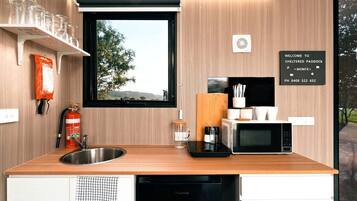 A compact kitchen with a sink, a microwave, and a toaster. There is a window with a view of trees and a sign that reads 'Welcome to Sheltered Paddock.'