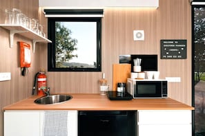 A compact kitchen with a sink, a microwave, and a toaster. There is a window with a view of trees and a sign that reads 'Welcome to Sheltered Paddock.'