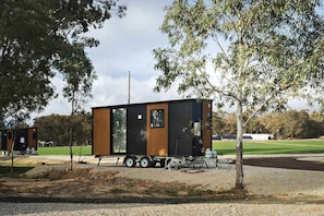 A modern, single-story mobile home with a flat roof, positioned on a gravel driveway with trees in the background.
