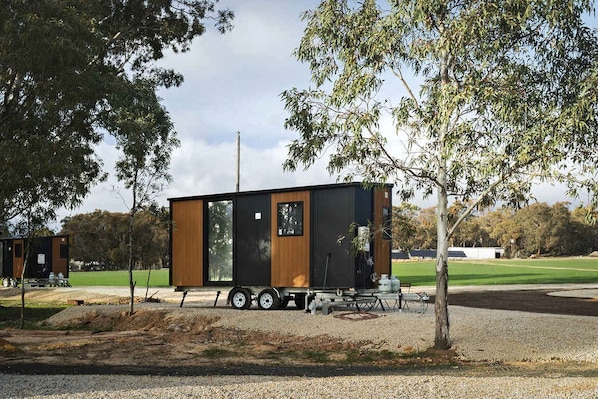 A modern, single-story mobile home with a flat roof, positioned on a gravel driveway with trees in the background.