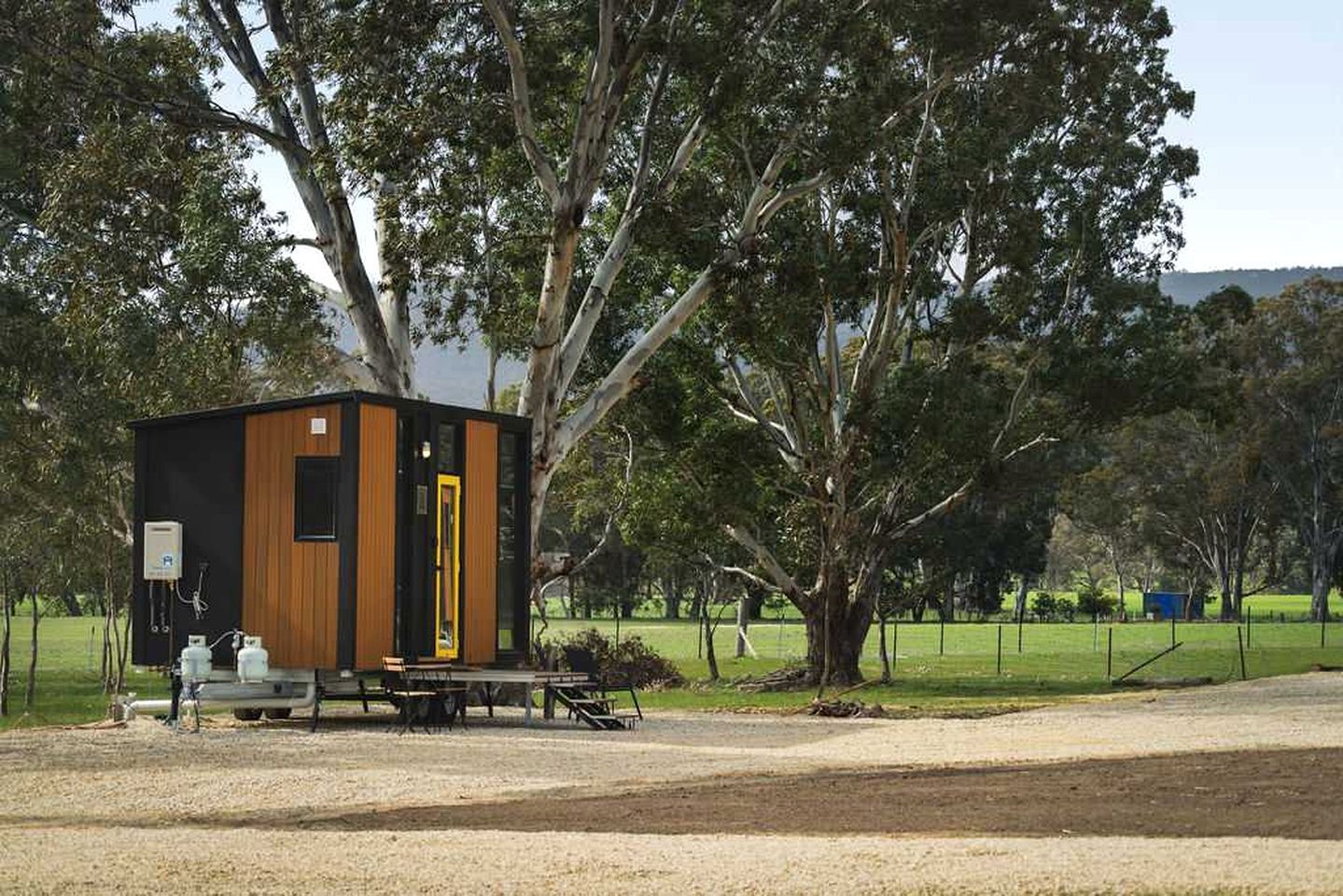 A small, single-story cabin with a wooden exterior and a yellow door, situated in a grassy area with trees and a fence in the background.
