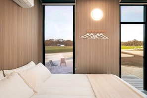 A bedroom with a bed, a view of a grassy area, and a wall-mounted light fixture.