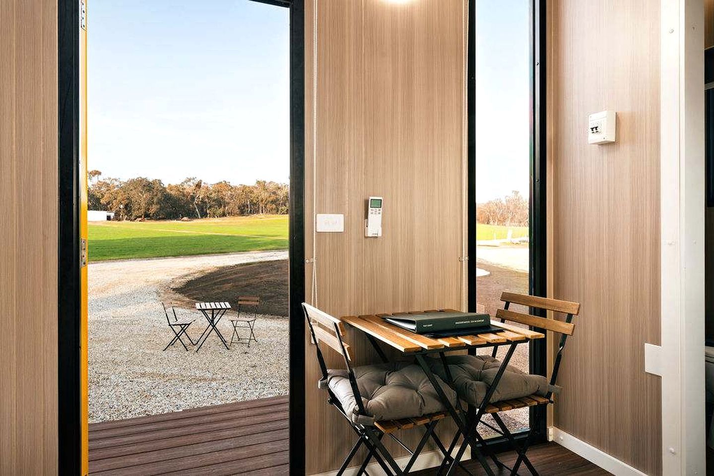 A room with a wooden interior, a table with chairs, a bench, and a view of a grassy area outside.