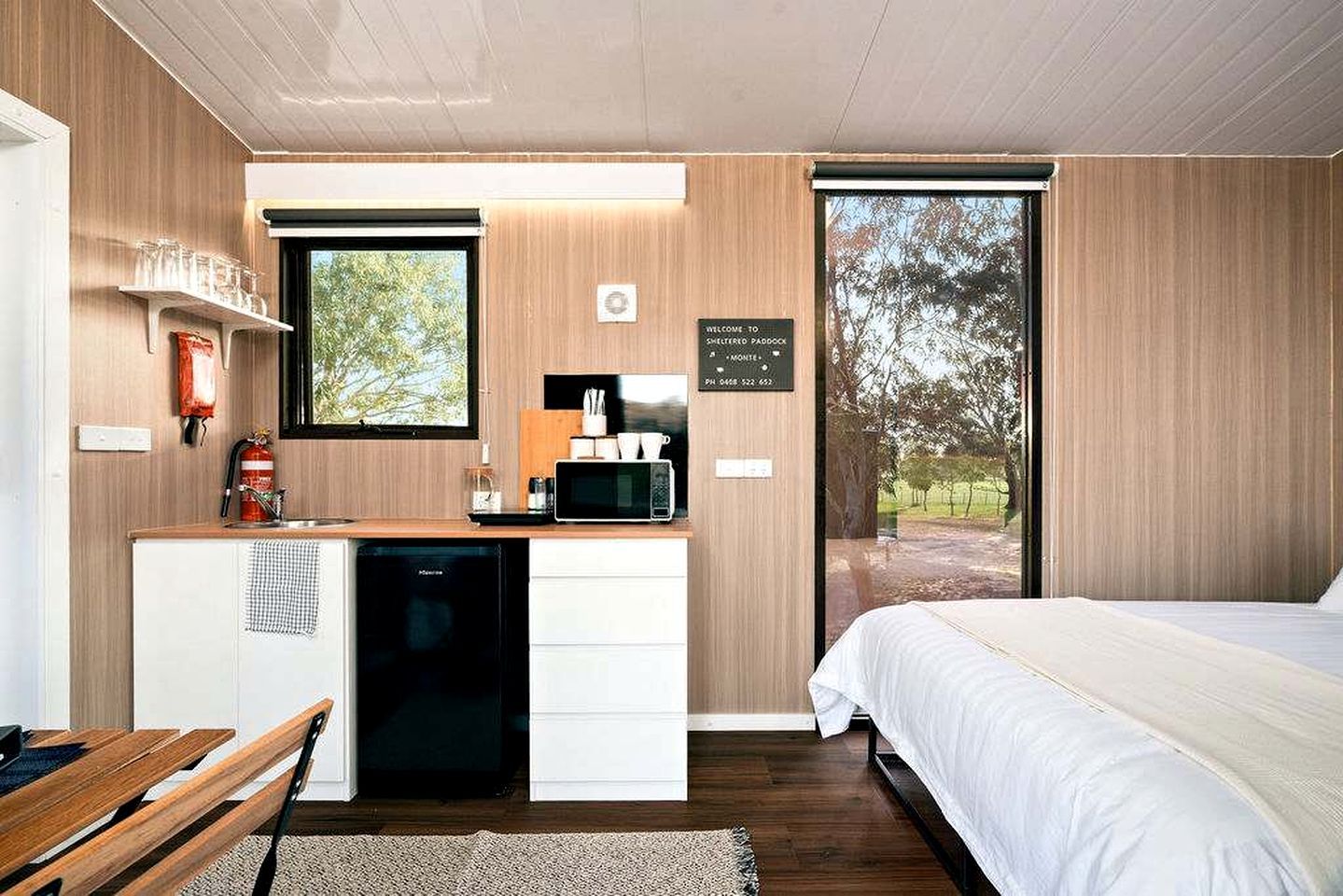 A modern bedroom with a bed, a desk with a microwave, a fire extinguisher, and a window with a view of trees.