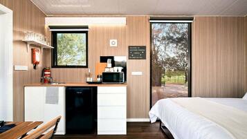 A modern bedroom with a bed, a desk with a microwave, a fire extinguisher, and a window with a view of trees.