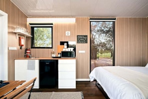 A modern bedroom with a bed, a desk with a microwave, a fire extinguisher, and a window with a view of trees.