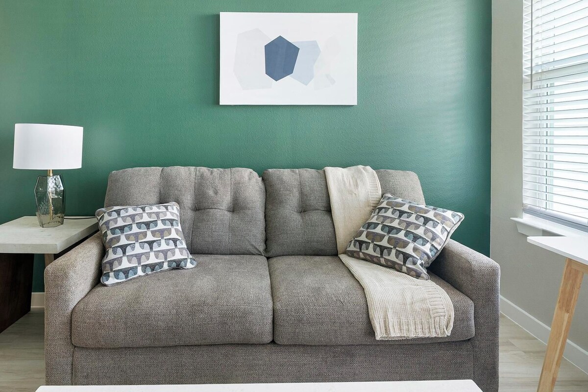 Un sofá gris con cojines estampados, una mesita auxiliar blanca con una lámpara y una obra de arte en una pared verde azulado.