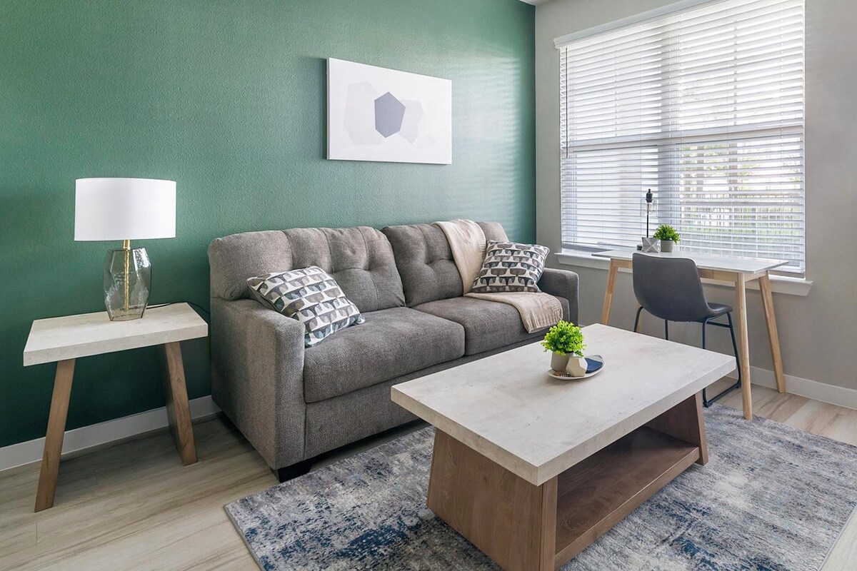 Una sala de estar con un sofá gris, una mesa de centro, una lámpara y una ventana con persianas.