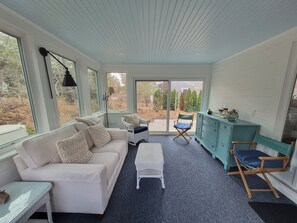 A room with a white sofa, a small table, a bench, a teal cabinet, and a view of the outdoors through large windows. - Charming Coastal Cottage near Cockle Cove Beach (S. Chatham)