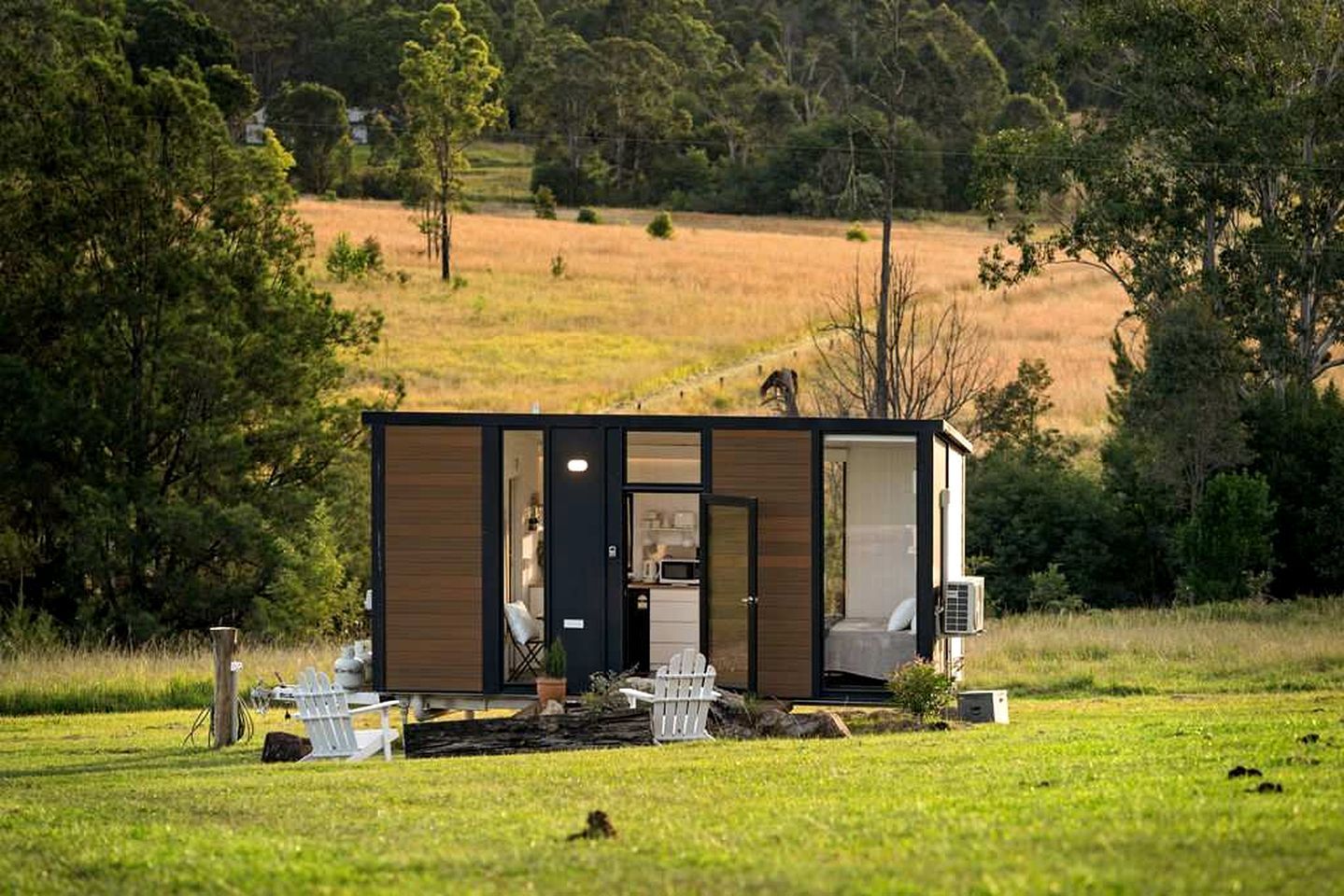 Une cabane moderne, de plain-pied, dotée d’une grande porte coulissante, d’un petit porche avec une table et des chaises, et offrant une vue sur un champ verdoyant et des arbres en arrière-plan.