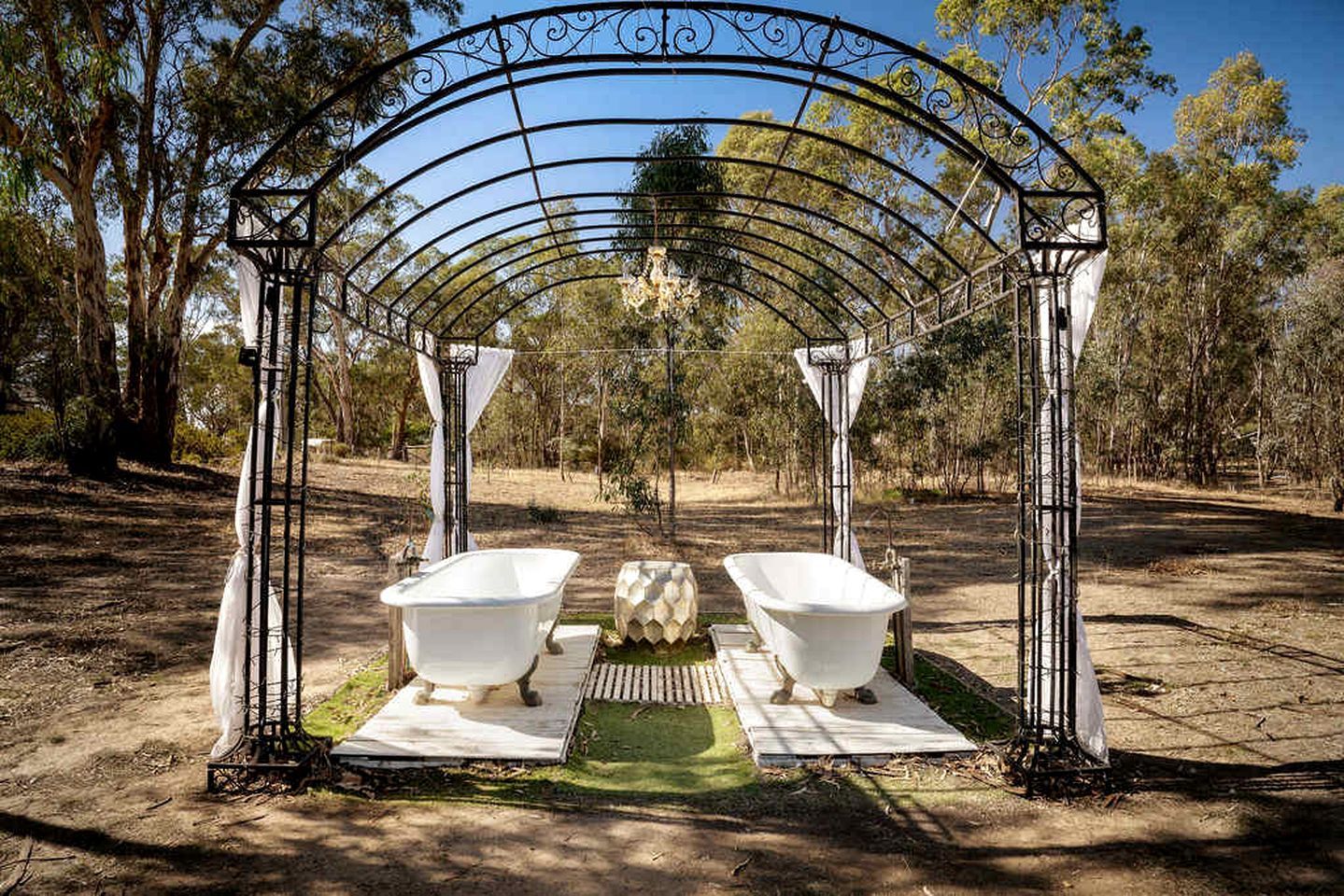 A metal archway with two white bathtubs under it, set in a natural outdoor area with trees.