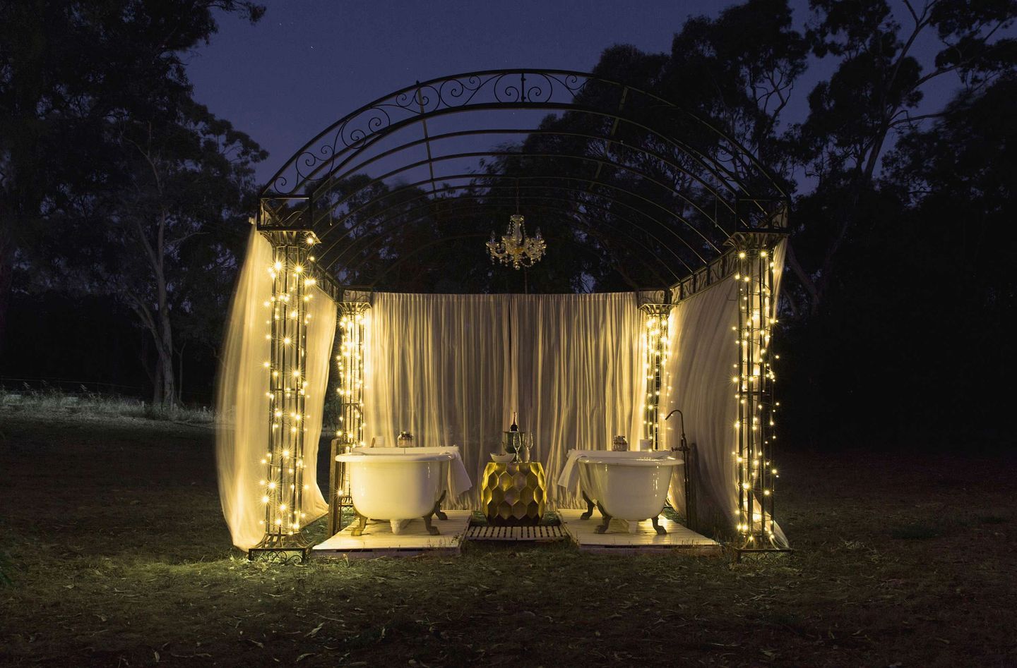 An outdoor gazebo with two white bathtubs, draped curtains, and string lights.