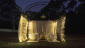 An outdoor gazebo with two white bathtubs, draped curtains, and string lights.