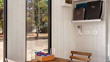 A room with a dining table and chairs, a bookshelf with books and a camera, and a view of trees through a large window.