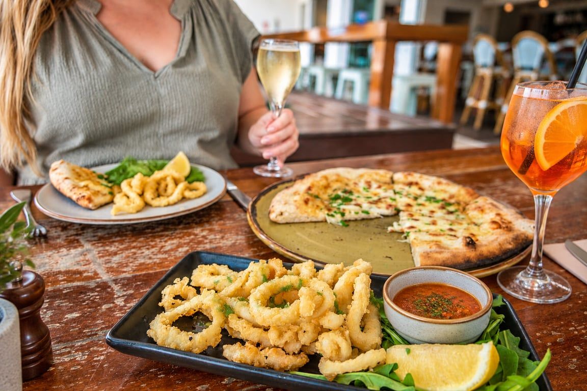 A dining table with a plate of fried calamari, a pizza, a glass of white wine, and a cocktail with an orange slice.
