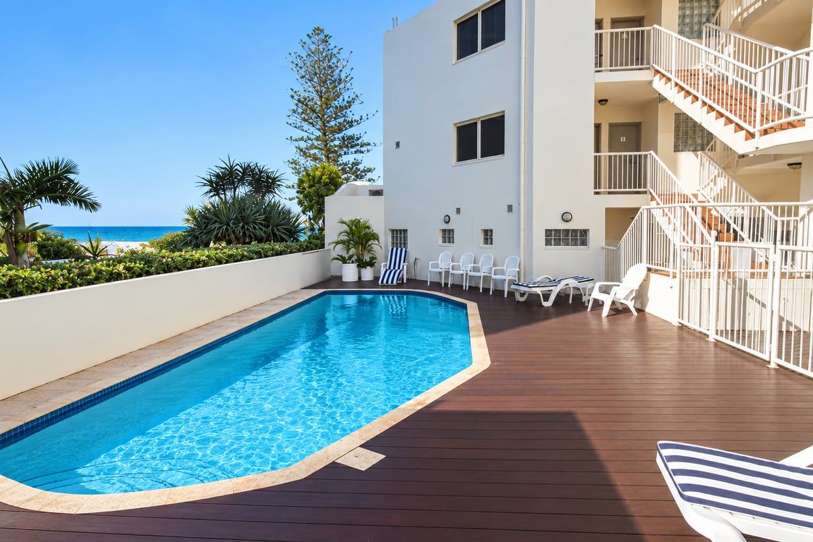 A swimming pool with a wooden deck, lounge chairs, and a view of the beach.