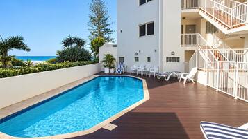 A swimming pool with a wooden deck, lounge chairs, and a view of the beach.