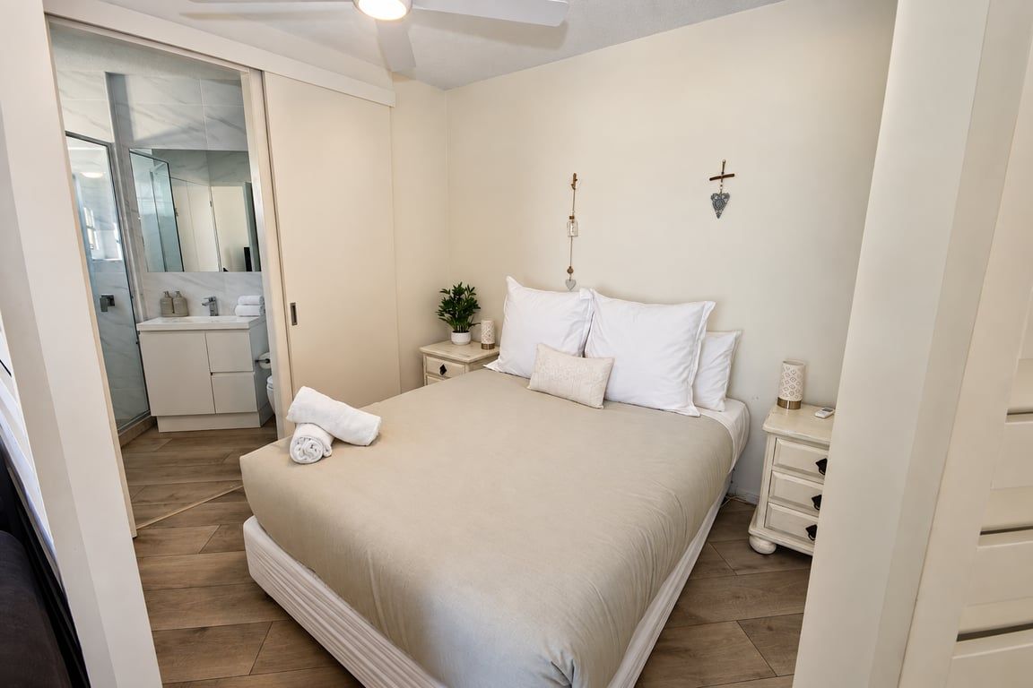 A bedroom with a bed, bedside tables, a nightstand, a wall-mounted light fixture, and a door leading to a bathroom.