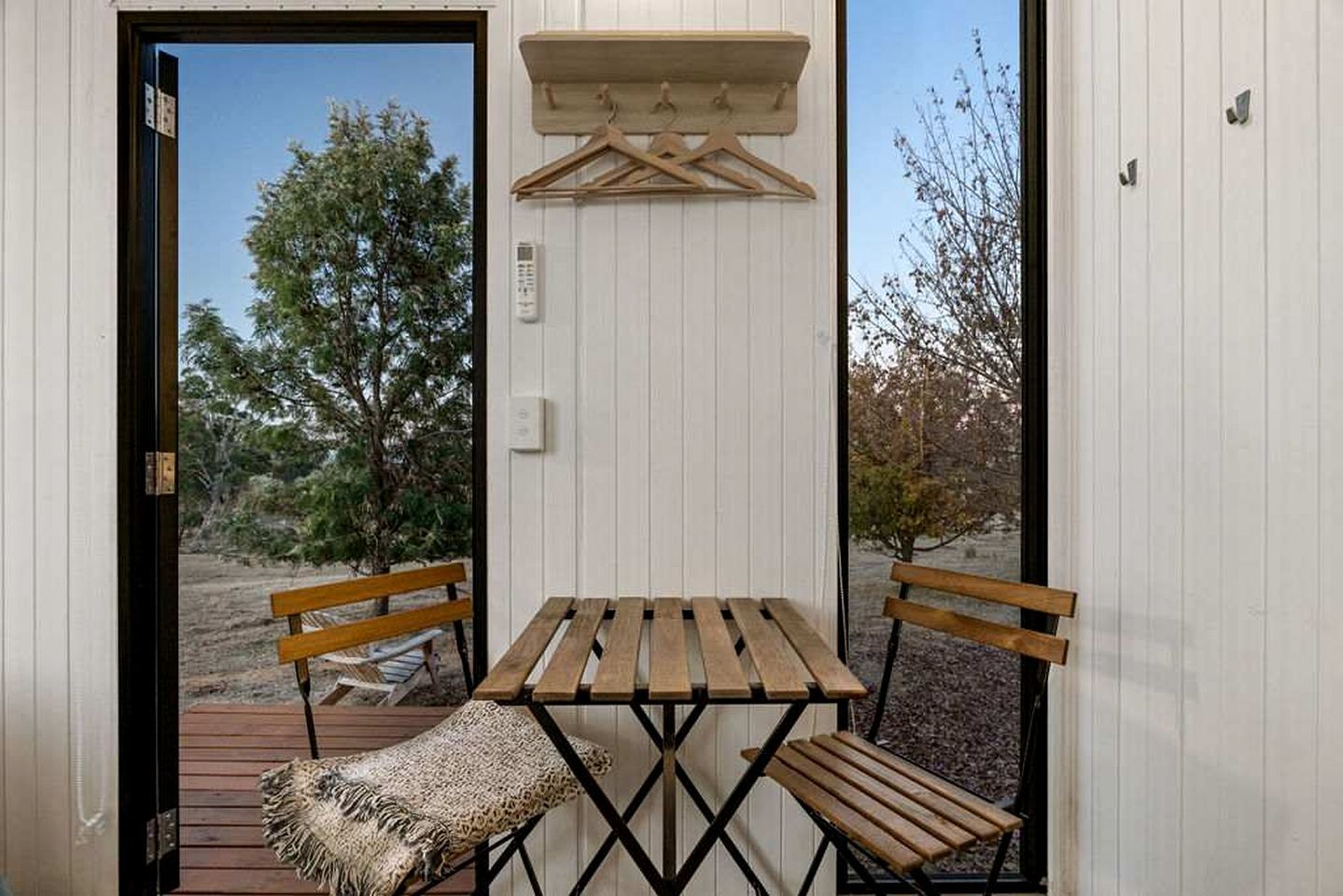 A small outdoor seating area with a wooden table and benches, a hanging coat rack, and a door leading outside.
