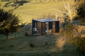 Een klein, modern vakantiehuisje met één deur en een zichtbare airconditioning, omringd door een grasveld en bomen.