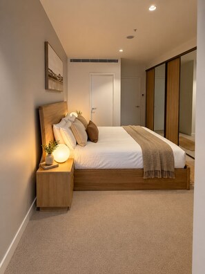 A bedroom with a wooden bed, bedside table, and a nightstand with a lamp. - Mocha Maison | Oversized 2 Bedroom, 2 Bathrooms + 2 Car Parks (Southbank)