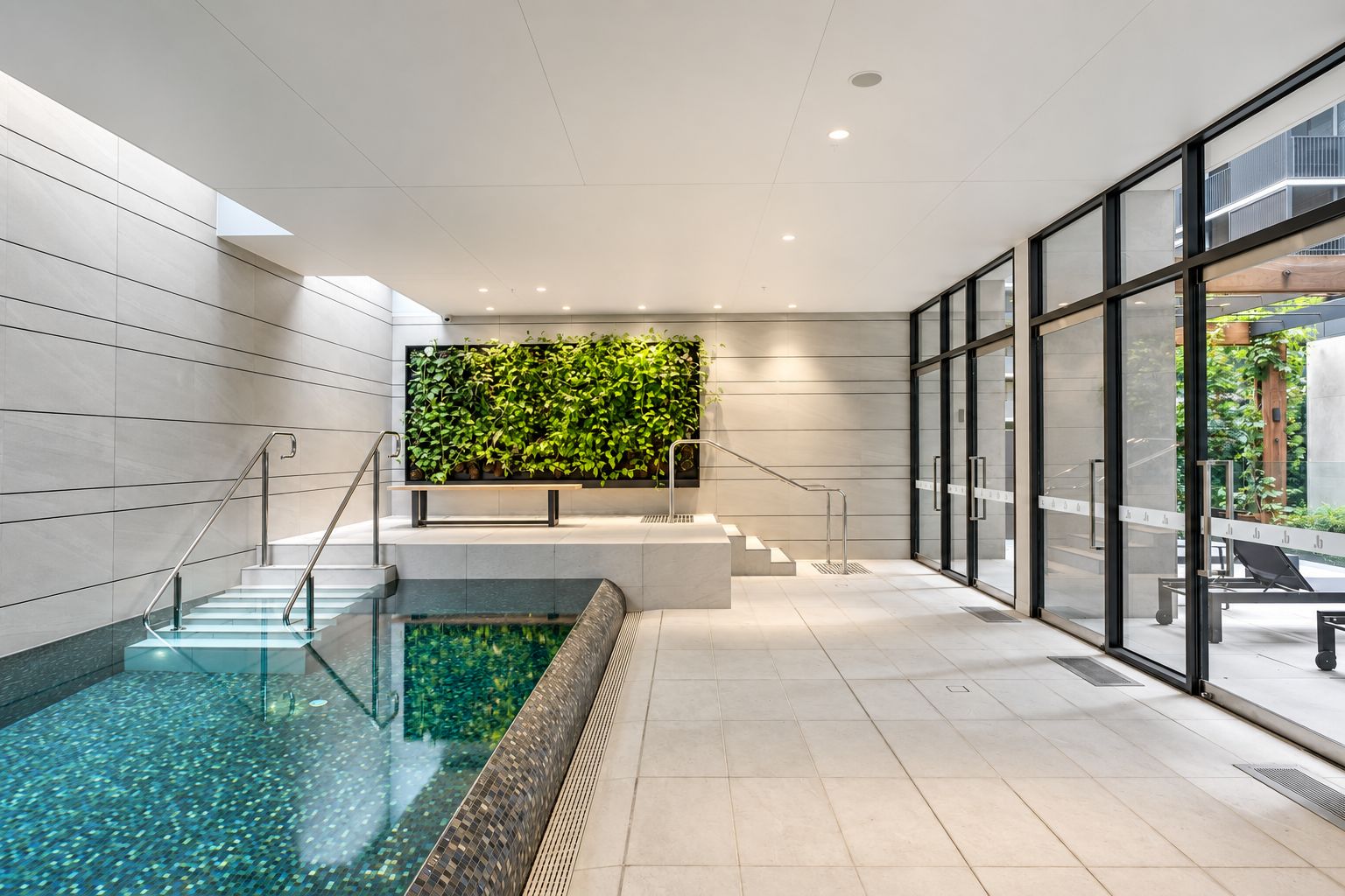 Un espace piscine intérieur moderne, doté d’un mur en verre, d’un escalier et d’un mur végétal.