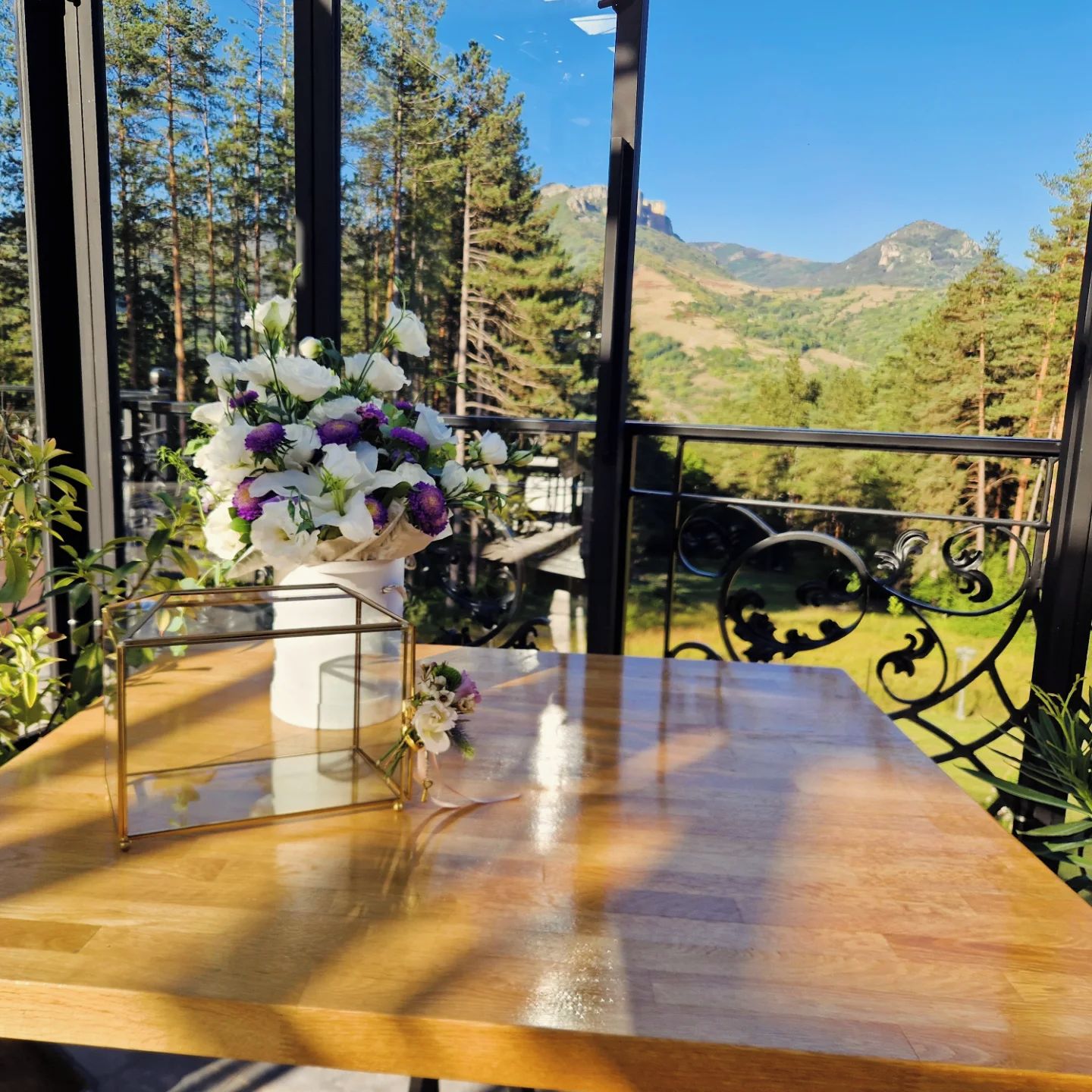 A wooden table with a vase of flowers and a glass container, set against a scenic mountain view.