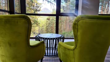 Een kamer met twee gele stoelen gericht op een ronde tafel, grote ramen met uitzicht op een bos en een berg.