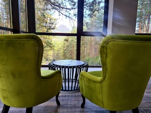 A room with two yellow chairs facing a round table, large windows with a view of a forest and a mountain.