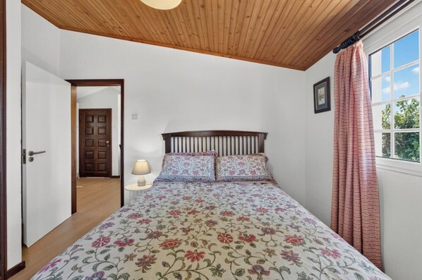 A bedroom with a wooden ceiling, a bed with floral bedding, a bedside lamp, and a window with curtains. - São Gonçalo View T1 (Funchal)
