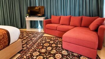 Quarto de hotel com cama, sofá vermelho, TV sobre suporte e tapete estampado.