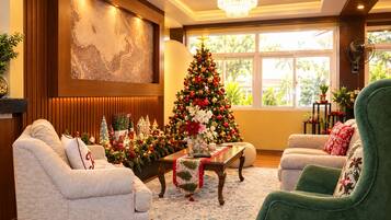 A cozy living room with a large Christmas tree, comfortable seating, and a view of greenery outside.