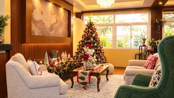 A cozy living room with a large Christmas tree, comfortable seating, and a view of greenery outside.