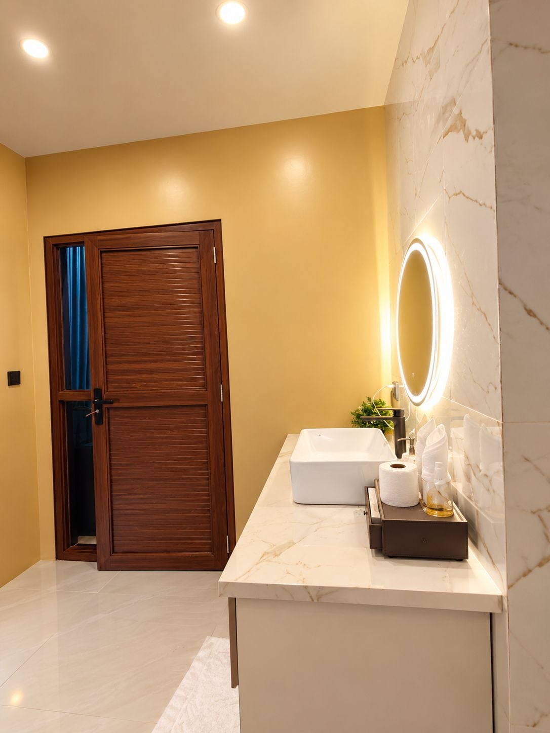 A modern bathroom with a marble vanity, a large mirror with lighting, a towel rack, and a wooden door.