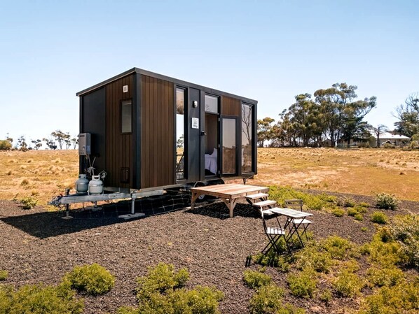 A modern, compact cabin with a wooden exterior and a sliding door, situated in a dry, grassy area with sparse vegetation.