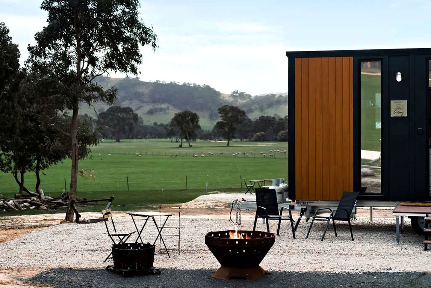 A modern outdoor seating area with a fire pit, chairs, and a view of a green field and trees.