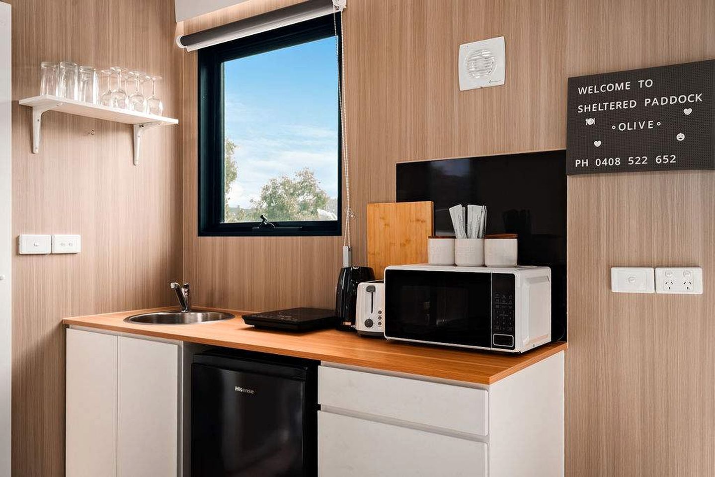A compact kitchen with a microwave, toaster, and a small sink. There's a window above the sink, and a welcome sign on the wall.