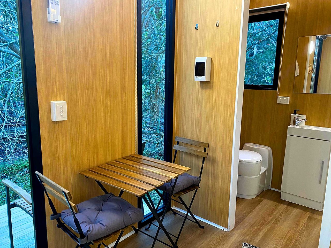 A compact room with a wooden table and two chairs, a small sink, and a window with a view of trees.